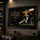 Caitlin Foord soccer wall art in a black frame displayed in a dark man cave with sports shelving and lounge seating.