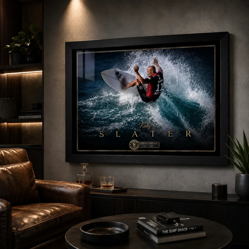Kelly Slater surfing wall art in a black frame displayed in a dark man cave with warm shelving and a leather chair.