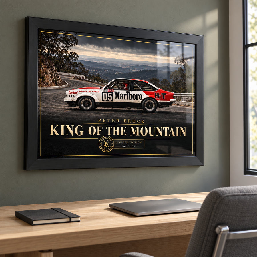 Black framed Peter Brock King of the Mountain wall art above a timber office desk beside a window.