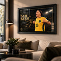 Black framed Sam Kerr Matildas soccer wall art displayed in a bright modern living room above a neutral sofa.