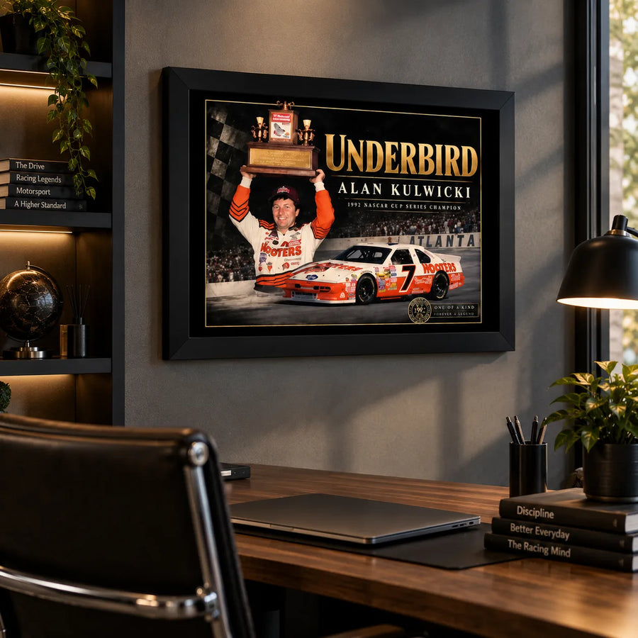 Black framed Alan Kulwicki Underbird wall art displayed above a wooden office desk with a task lamp.